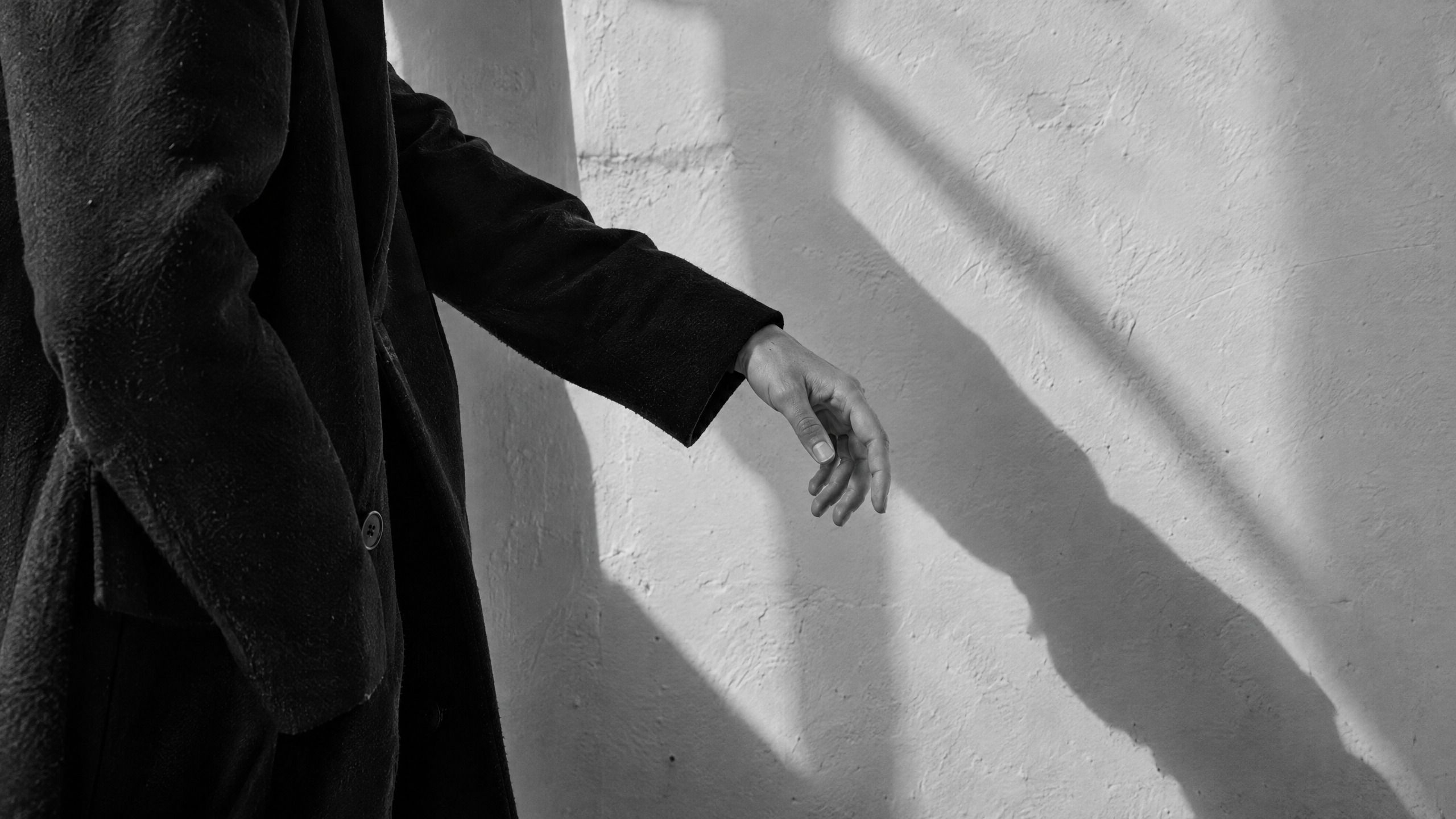 Die Hand eines Menschen im dunklen Mantel ruht vor einer verputzten Wand. Das Fensterlicht zeichnet einen einzelnen klaren Schatten.